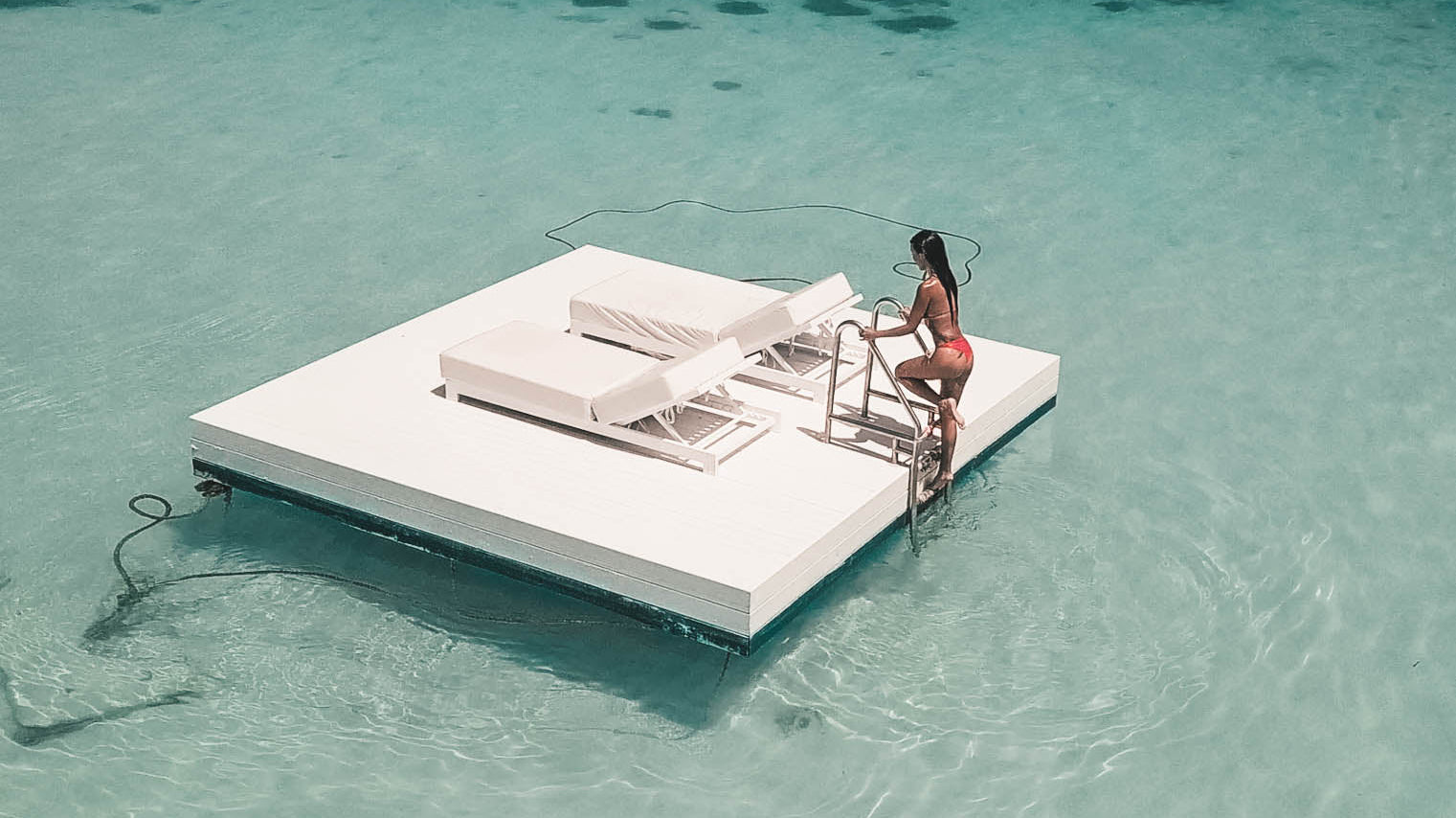 A woman in a swimsuit poses confidently on a contemporary floating pool deck with pristine white sun loungers, surrounded by translucent turquoise water.