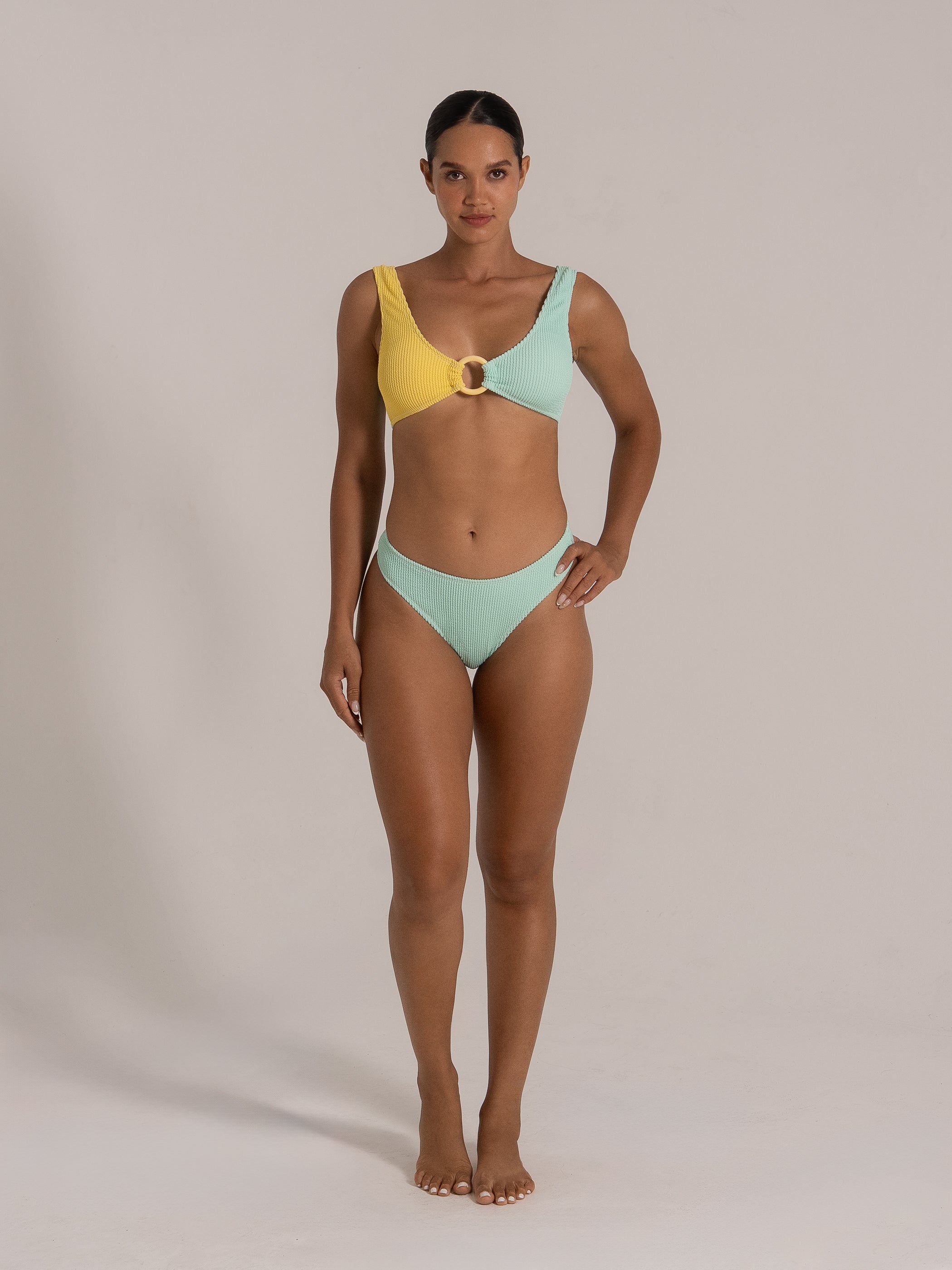 Front view of a model wearing a yellow and mint green colorblock bikini top with a central ring detail and matching mint bottom.
