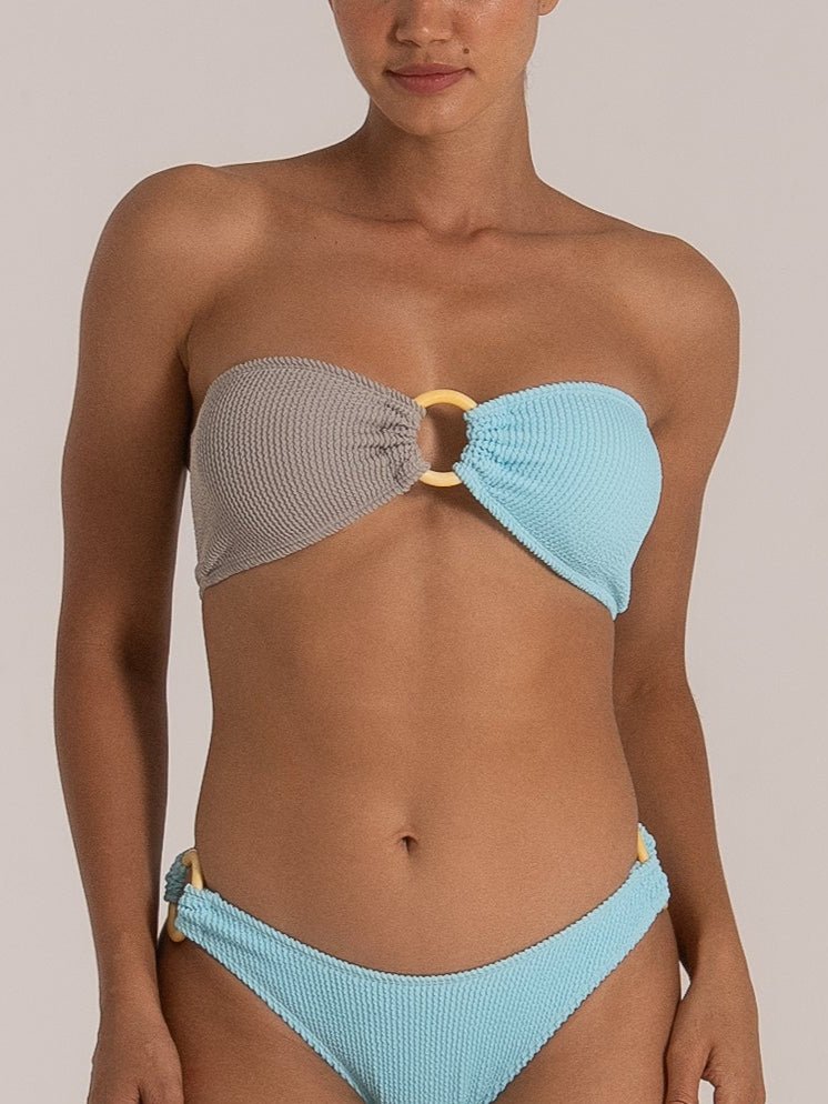 Close-up front view of model wearing a grey and blue bandeau bikini with central ring detail and high-cut textured bottoms.