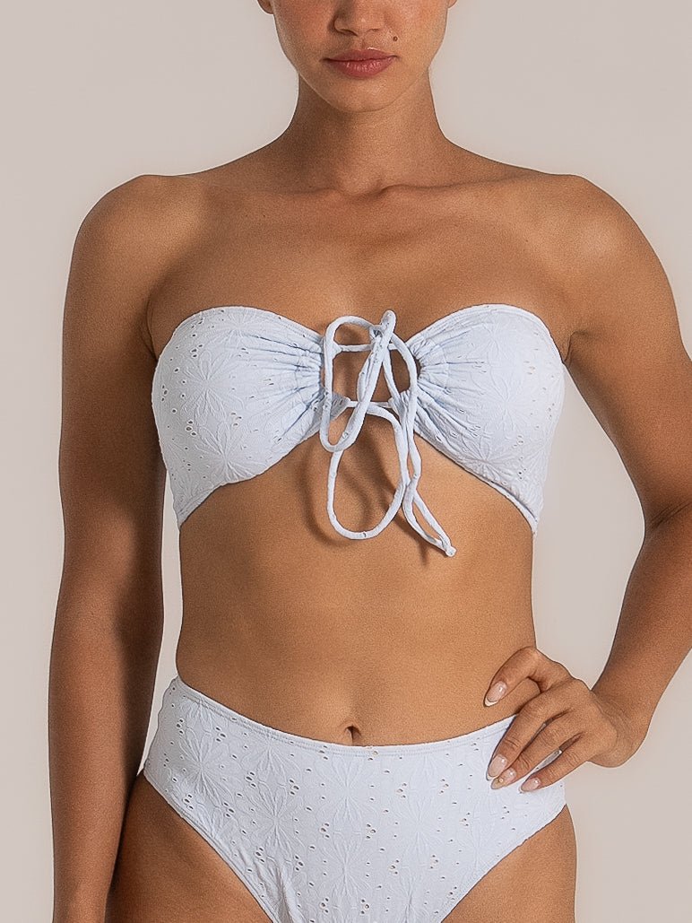 Front view of a woman wearing a light blue embroidered bandeau bikini set featuring eyelet floral patterns, a ruched front with tie detail, and high-waisted bottoms.