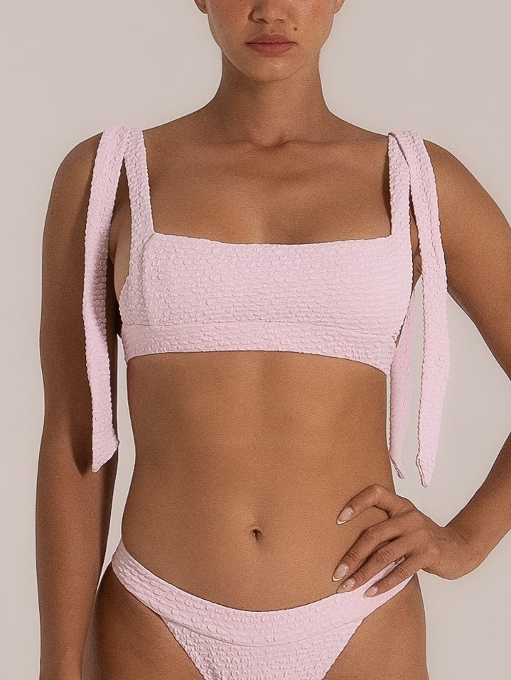 Front view of a woman wearing a light pink textured bikini set featuring wide self-tie shoulder straps and a square neckline, paired with a matching V-cut bottom for a feminine and flattering silhouette.