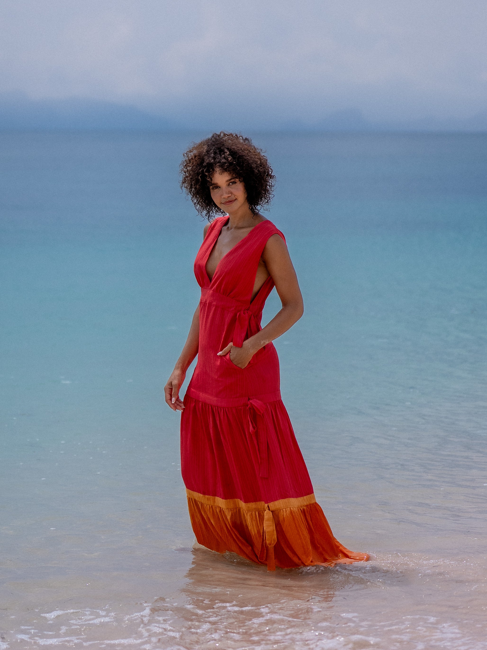 Woman standing in the ocean wearing OCEAN MYSTERY red sleeveless maxi dress with orange ombre hem.