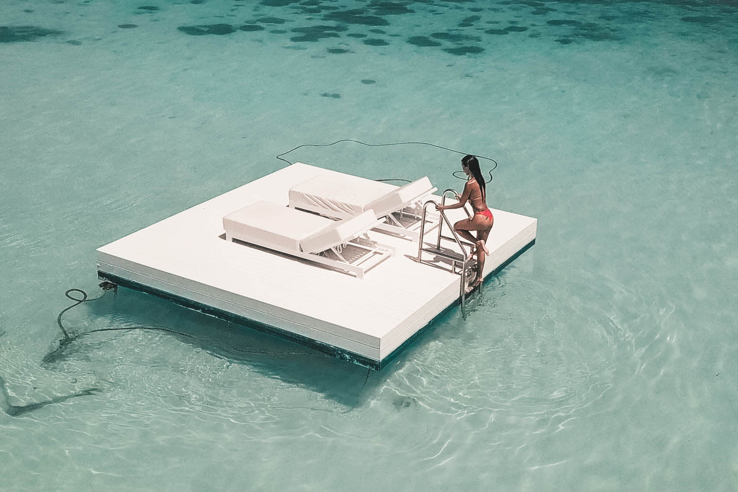 A woman in a swimsuit poses confidently on a contemporary floating pool deck with pristine white sun loungers, surrounded by translucent turquoise water.