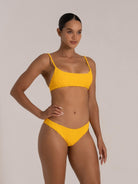 A fit model wearing a textured yellow two - piece bikini with thin straps. She has her hand on her hip, standing against a plain background.