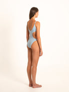 Slight back angle of model in textured blue monokini with waist cutout and soft straps