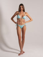 Full body shot of a model in an aqua shimmer triangle bikini with halter ties and side-tie bottoms, standing with hands on hips.