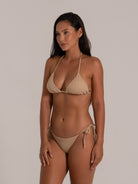 Woman in beige triangle bikini with halter tie and string sides, posing at a side angle in stud