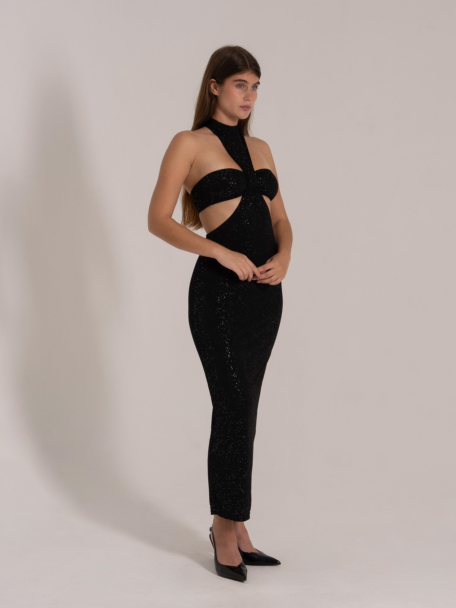 Side angle of a fitted black glitter halter dress with front cutout and high neck detail.