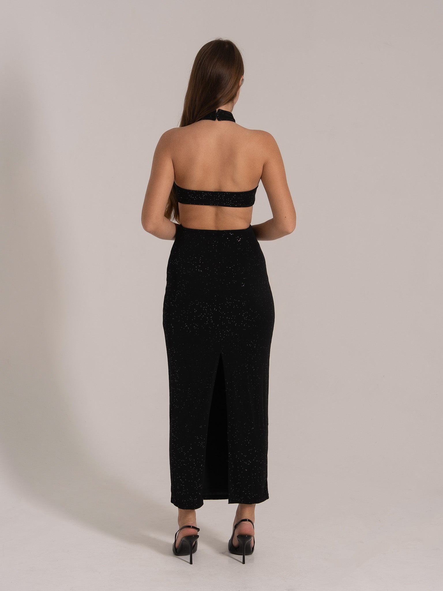 Back view of a woman wearing a black halter-neck cutout sparkle maxi dress with an open back and back slit.