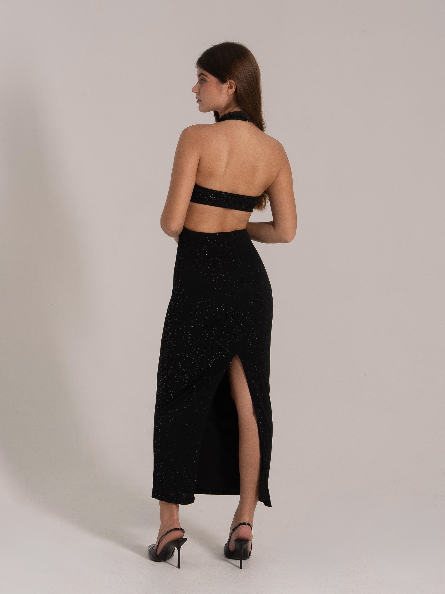 Woman in black sparkle halter evening dress with cutout back and thigh slit, side-back angle.