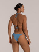 Woman wearing a blue textured triangle bikini from the back, showcasing tie straps and minimal coverage style.