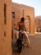 Resort lifestyle shot of a woman on a vintage scooter in a Moroccan-style courtyard, wearing a charcoal keyhole swimsuit styled as a bodysuit with loose white trousers.