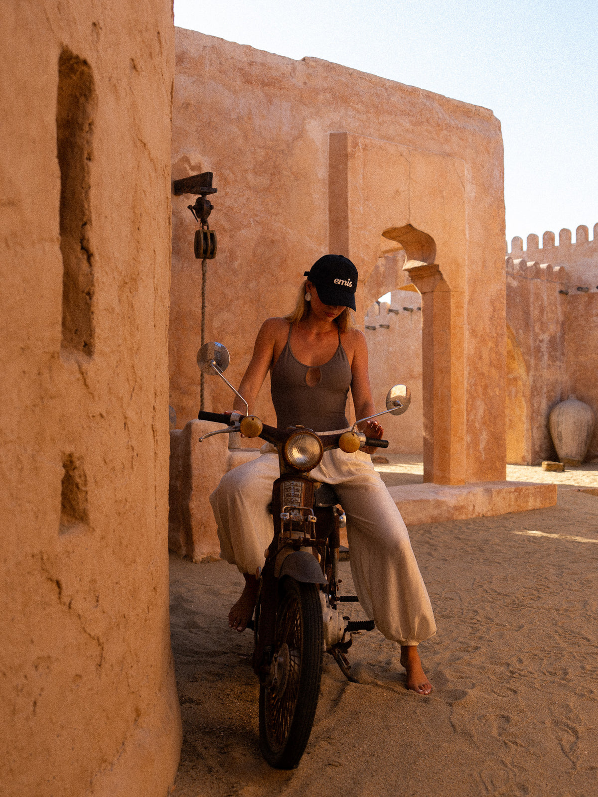 Resort lifestyle shot of a woman on a vintage scooter in a Moroccan-style courtyard, wearing a charcoal keyhole swimsuit styled as a bodysuit with loose white trousers.