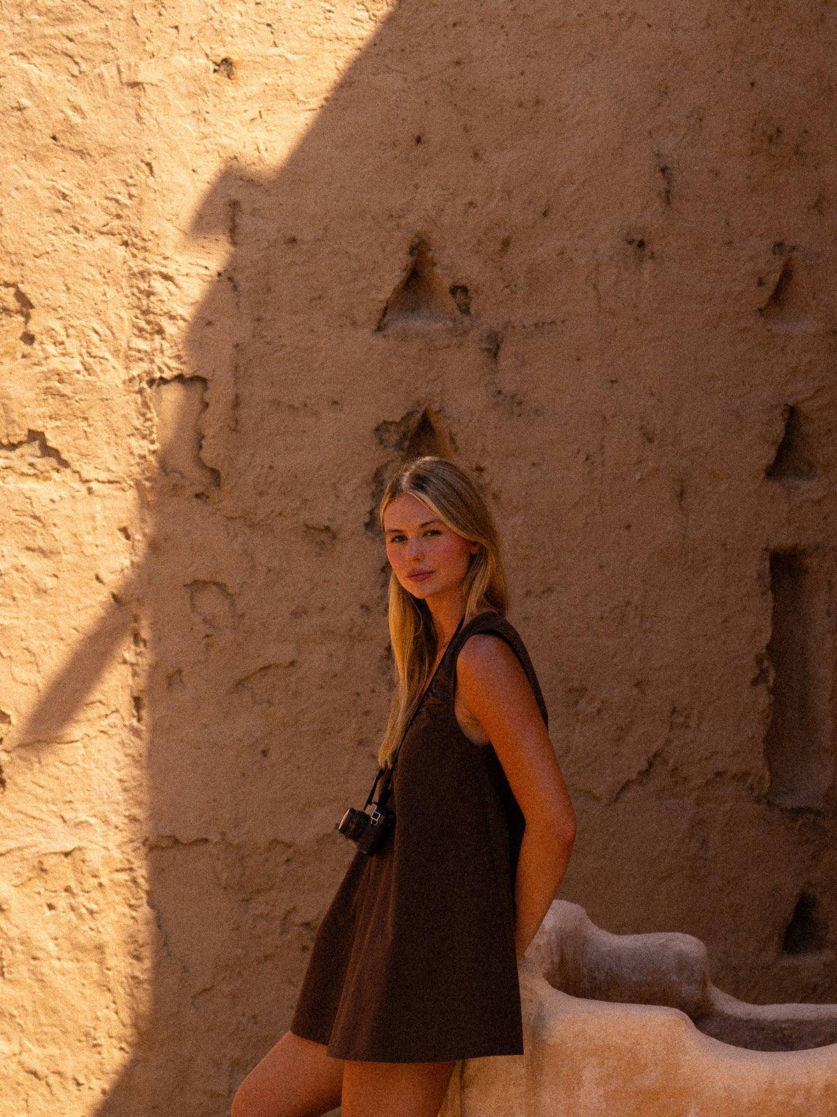 A woman modeling the chocolate brown ruched V-neck mini dress and a vintage camera against a textured mud-brick wall for a chic resort travel aesthetic.