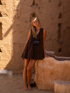 Lifestyle shot of a woman wearing a chocolate brown reversible cotton mini dress, carrying a vintage camera while leaning against a sunlit terracotta wall.