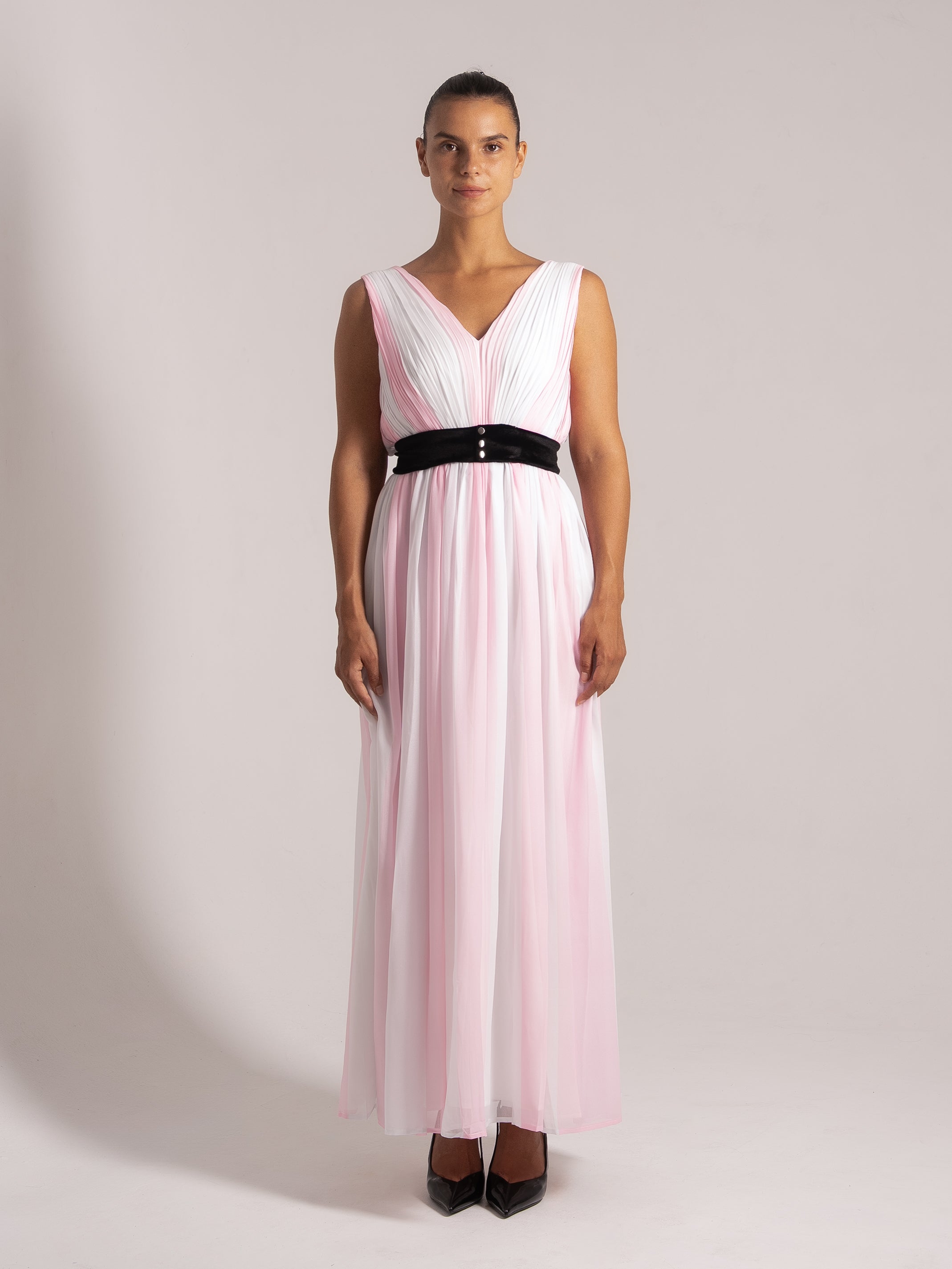 Woman wearing a pleated ombre pink and white maxi dress with a deep V-neck and a black velvet waistband, front view.