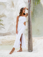 Elegant woman in a white one-shoulder long dress with a waist cut-out, leaning against a palm tree.