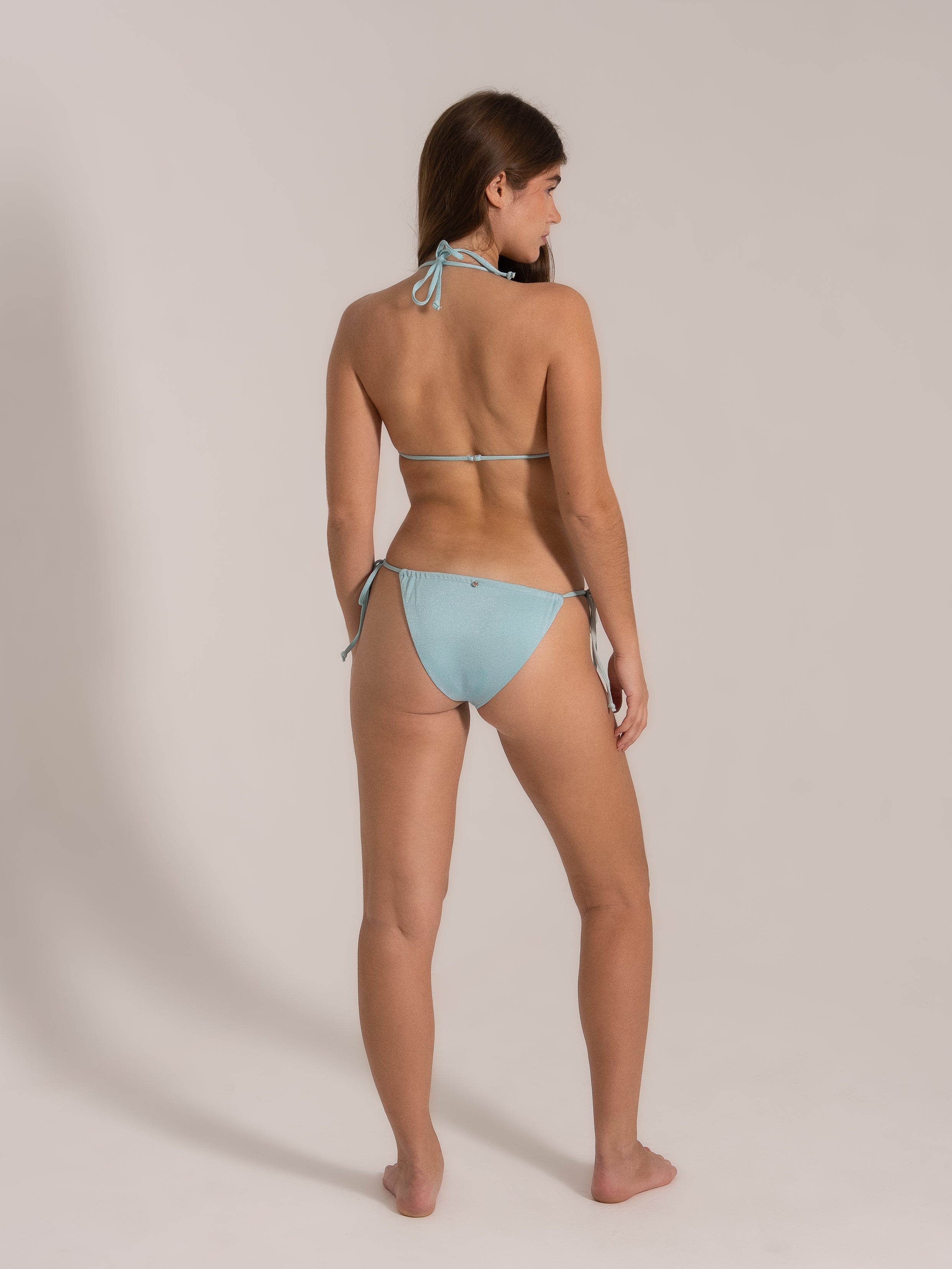 Model posing in a light blue string bikini set, side-back angle highlighting tie straps and cheeky fit