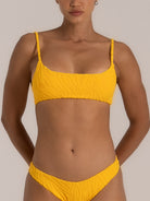 Front close-up of a woman wearing a mustard yellow textured bikini set featuring a bandeau-style top with braided straps and matching mid-rise bottoms.