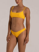 Side angle view of a woman in a mustard yellow textured bikini featuring braided straps and seamless edges.