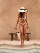 Woman walking by a pool wearing OCEAN MYSTERY pink gingham tie-front bikini set with spaghetti straps and a large straw hat.