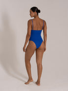 Woman wearing a deep blue one-piece swimsuit with textured fabric, low back and adjustable spaghetti straps.