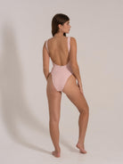 Back side view of a model in a pastel pink one-piece swimsuit with ribbed texture and low cut back.