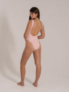 Woman wearing a pastel pink textured one-piece swimsuit with a low scoop back, posing from the back.
