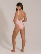 Woman wearing a light pink one-piece swimsuit with a low scoop back, standing and looking over shoulder
