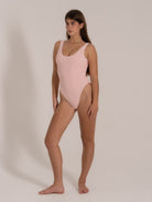 Female model in a pastel pink one-piece swimsuit posing in a side angle with high-leg cut