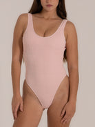 Front view of a woman wearing a light pink textured one-piece swimsuit with a scoop neckline and high-cut leg design, offering a soft and feminine silhouette.