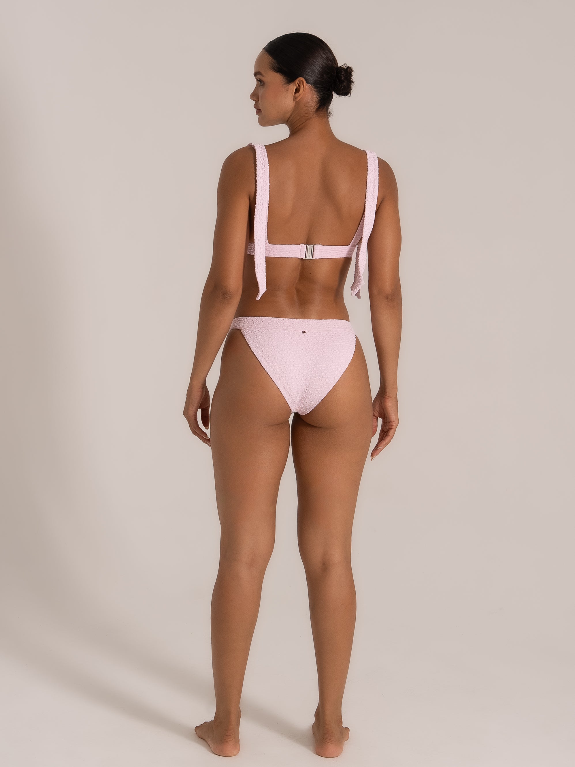 Back view of model Kate wearing a light pink textured bikini with wide shoulder ties and clasp back.