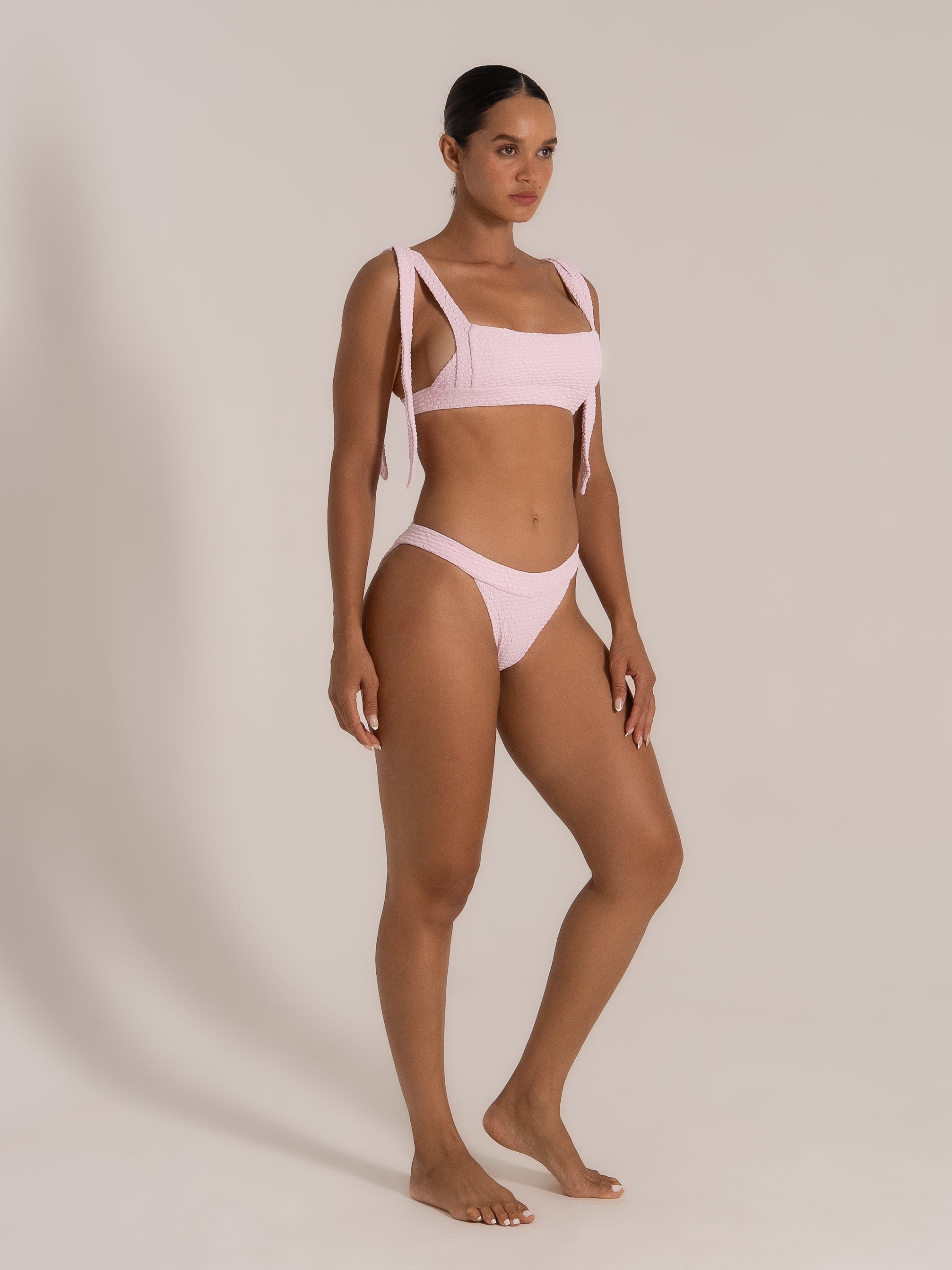 Model posing in a pastel pink bikini with bow-tie shoulder straps and high-leg bottoms.