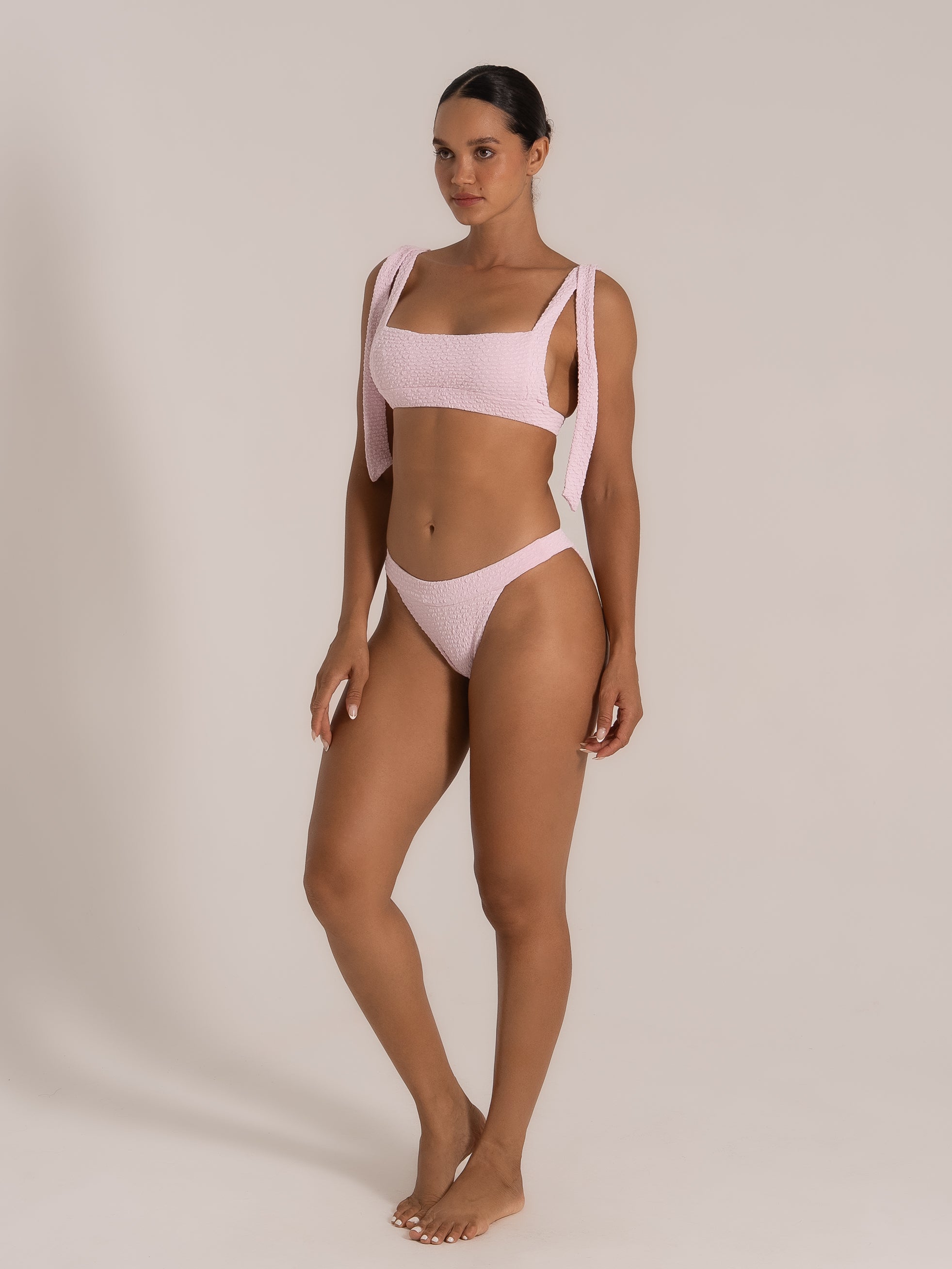 Front right view of model in a pink crinkled bikini with thick tie-up shoulder straps.