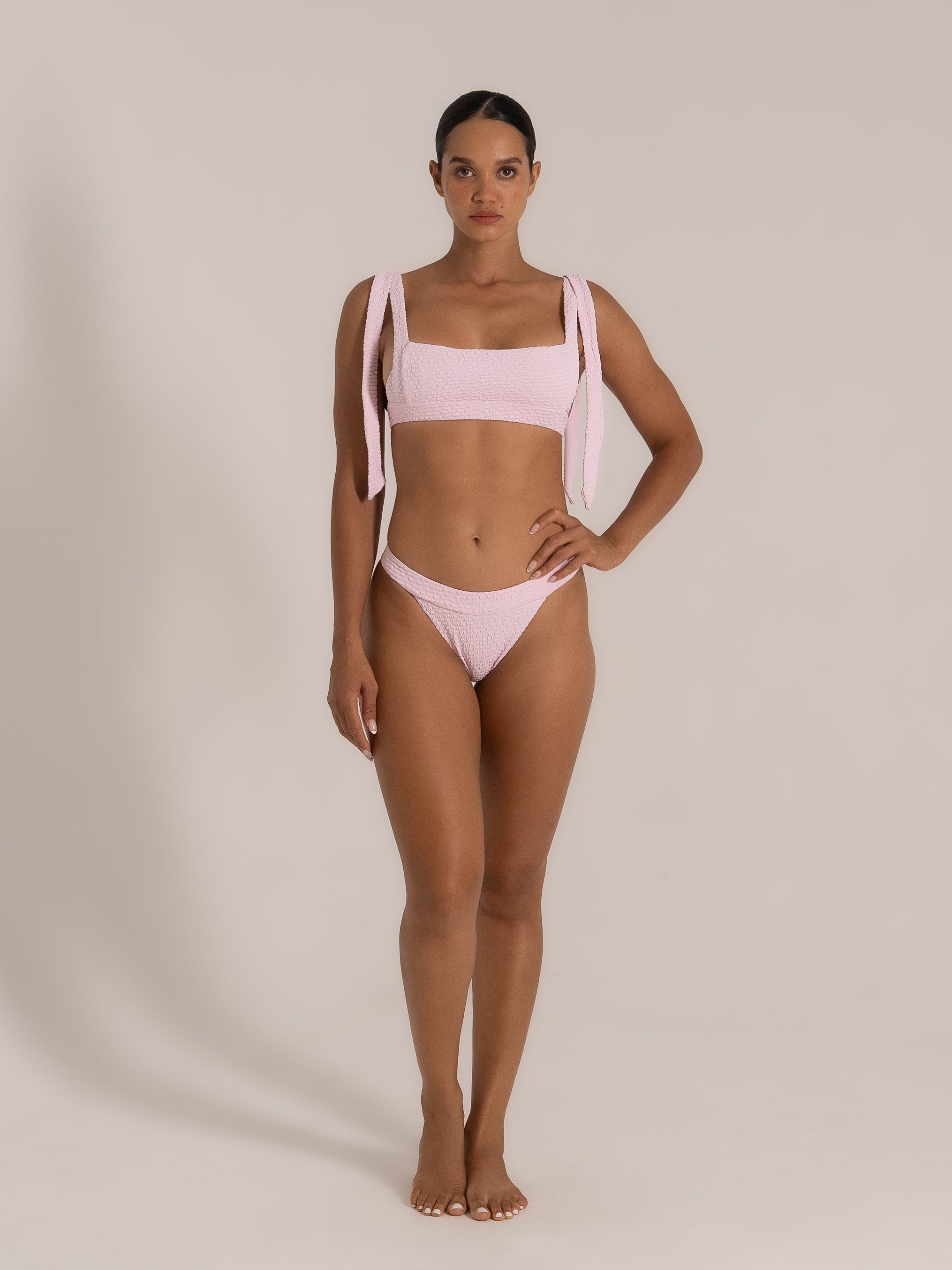 Full front view model wearing a soft pink textured bikini featuring wide tied straps and high-cut bikini bottom.