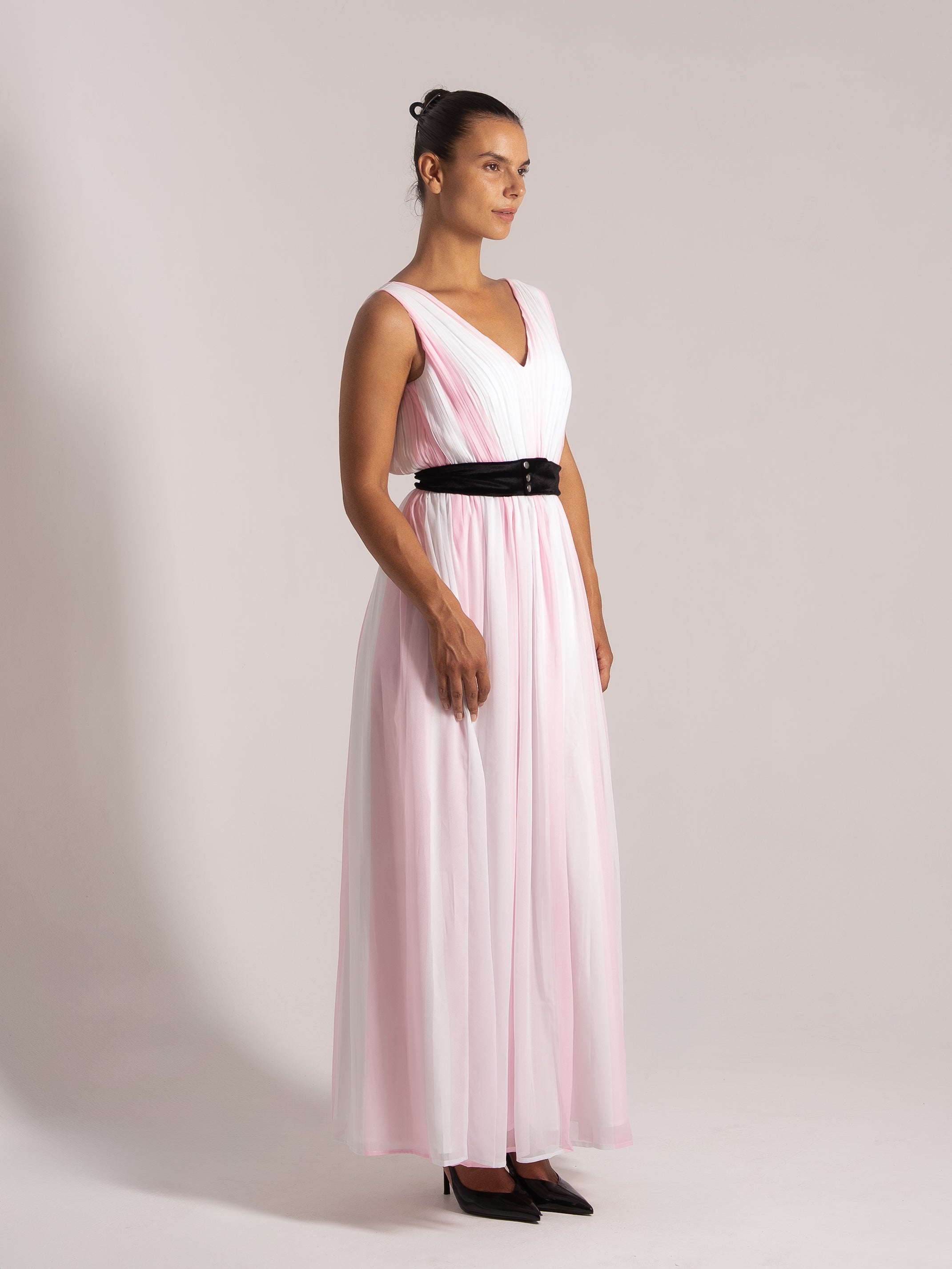 Side angle of a woman modeling a pink and white pleated maxi dress with a flattering silhouette and black belt detail.