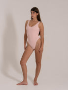 Model in a pastel pink scoop-neck one-piece swimsuit, showing a flattering side angle with textured fabric.