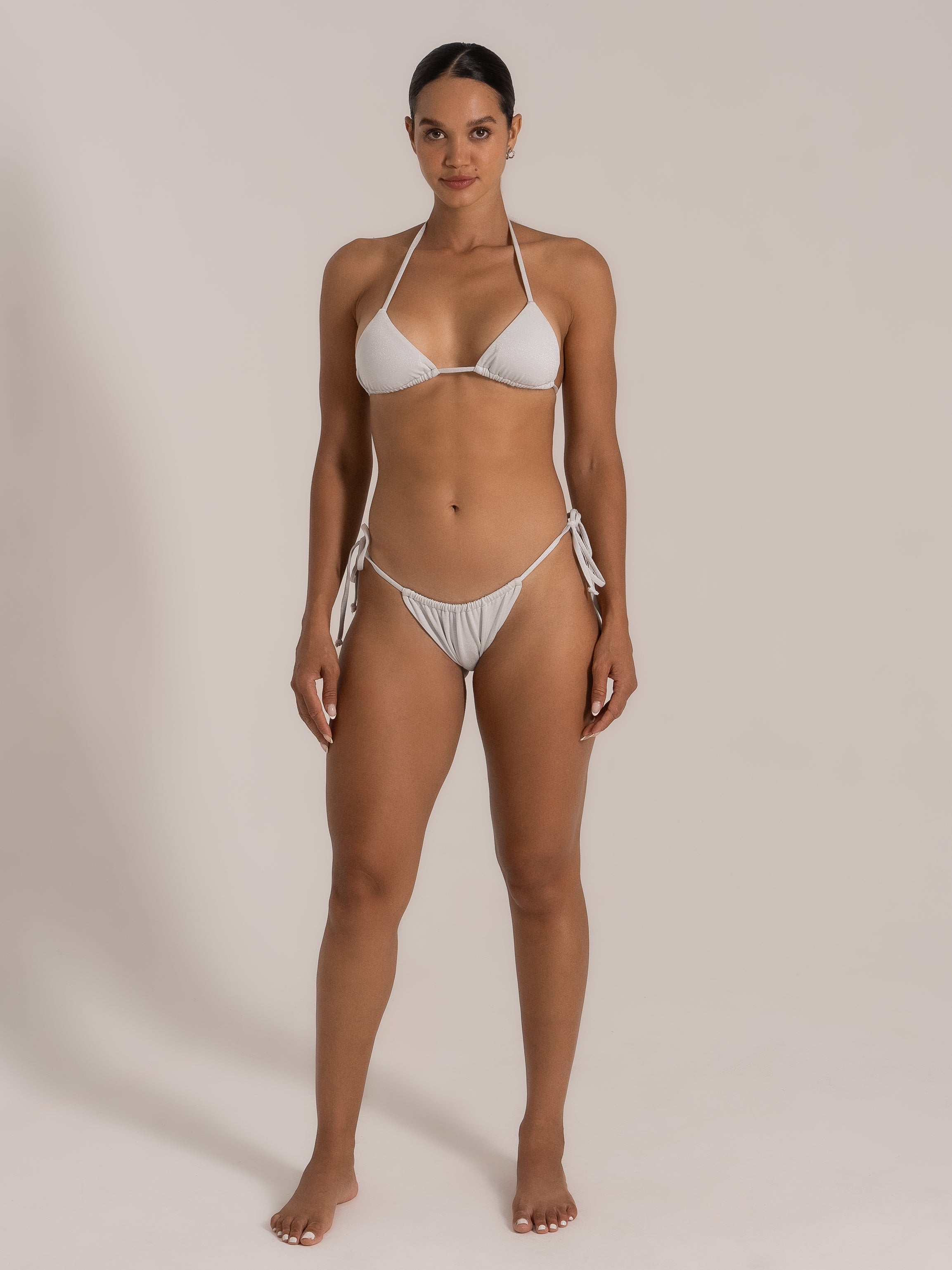 Front view of model in white shimmer triangle bikini set with halter tie and ruched bottoms.