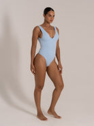 Model posing in a baby blue V-neck one-piece swimsuit with textured fabric and angled stance.
