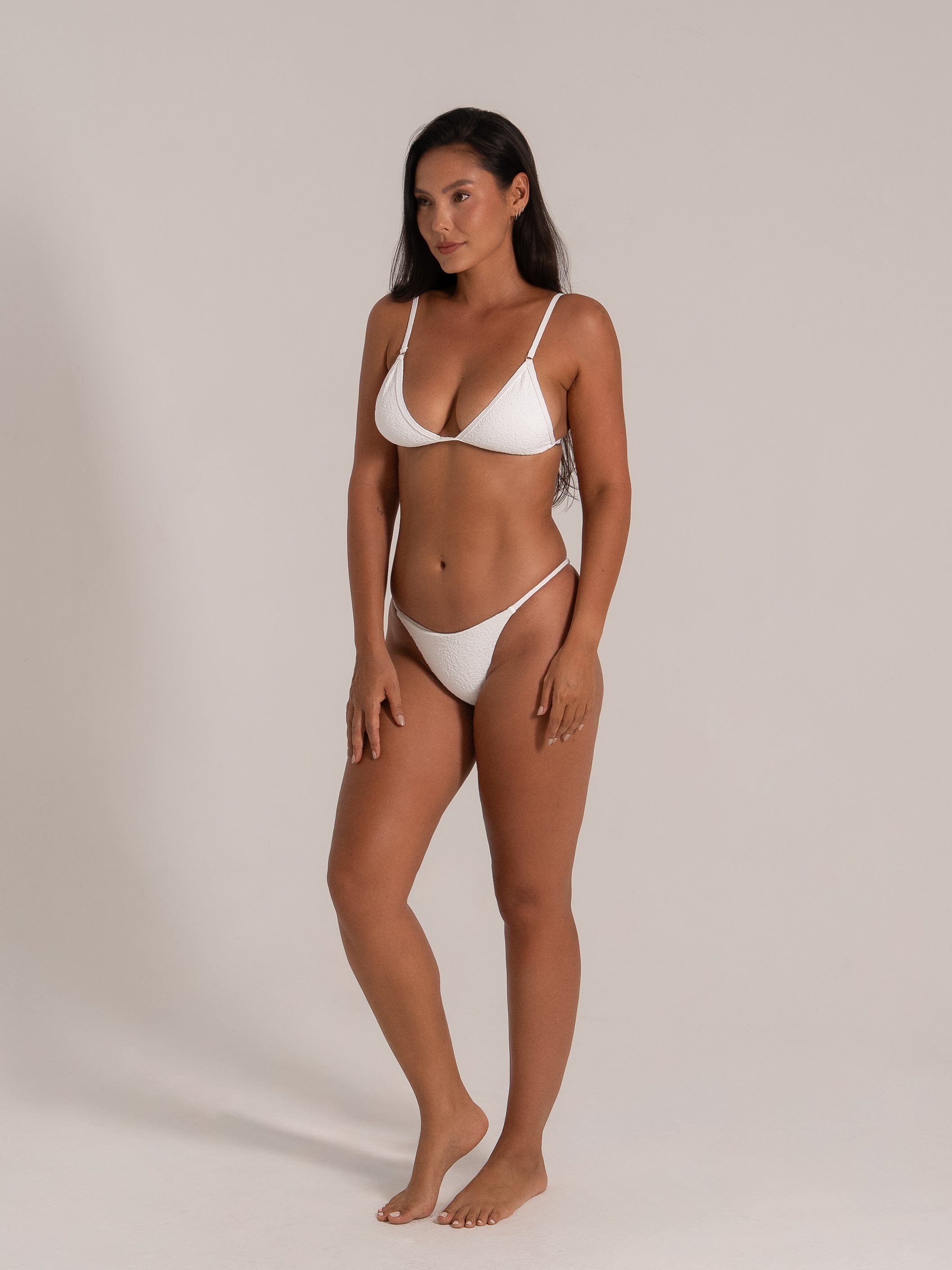 Model posing in white bikini set with slim side straps and textured detailing, perfect for a sleek beach look.