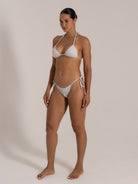Side pose of model in white metallic triangle bikini featuring adjustable halter and side ties.