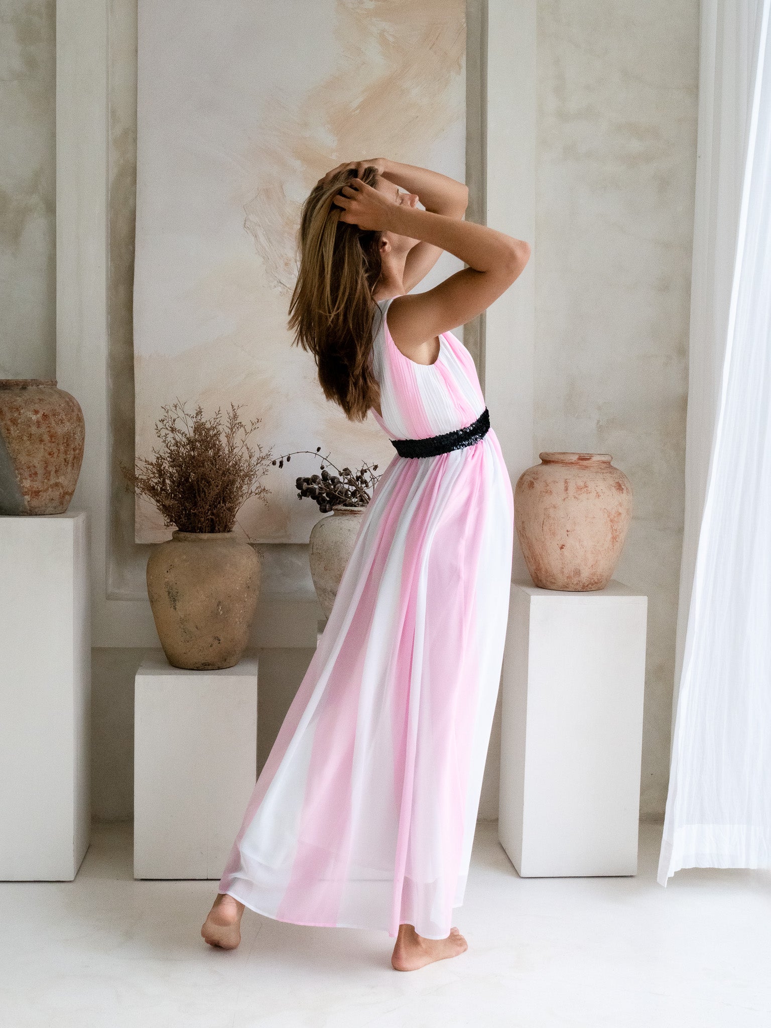 Woman wearing a V-neck white and pink pleated maxi dress with a black sequin waist belt.