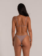 A woman seen from the back wearing a light - purple lingerie set, including a thin - strapped top and a thong bottom, showcasing her tanned and fit physique against a neutral background.