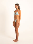Model in wrap-front light blue bikini, posing at a soft right angle with minimal backdrop