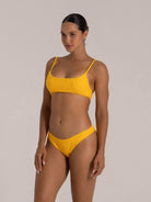 A confident model showcases a vibrant yellow textured bikini with thin straps and low-rise briefs, highlighting her toned midriff and fit physique against a neutral backdrop.