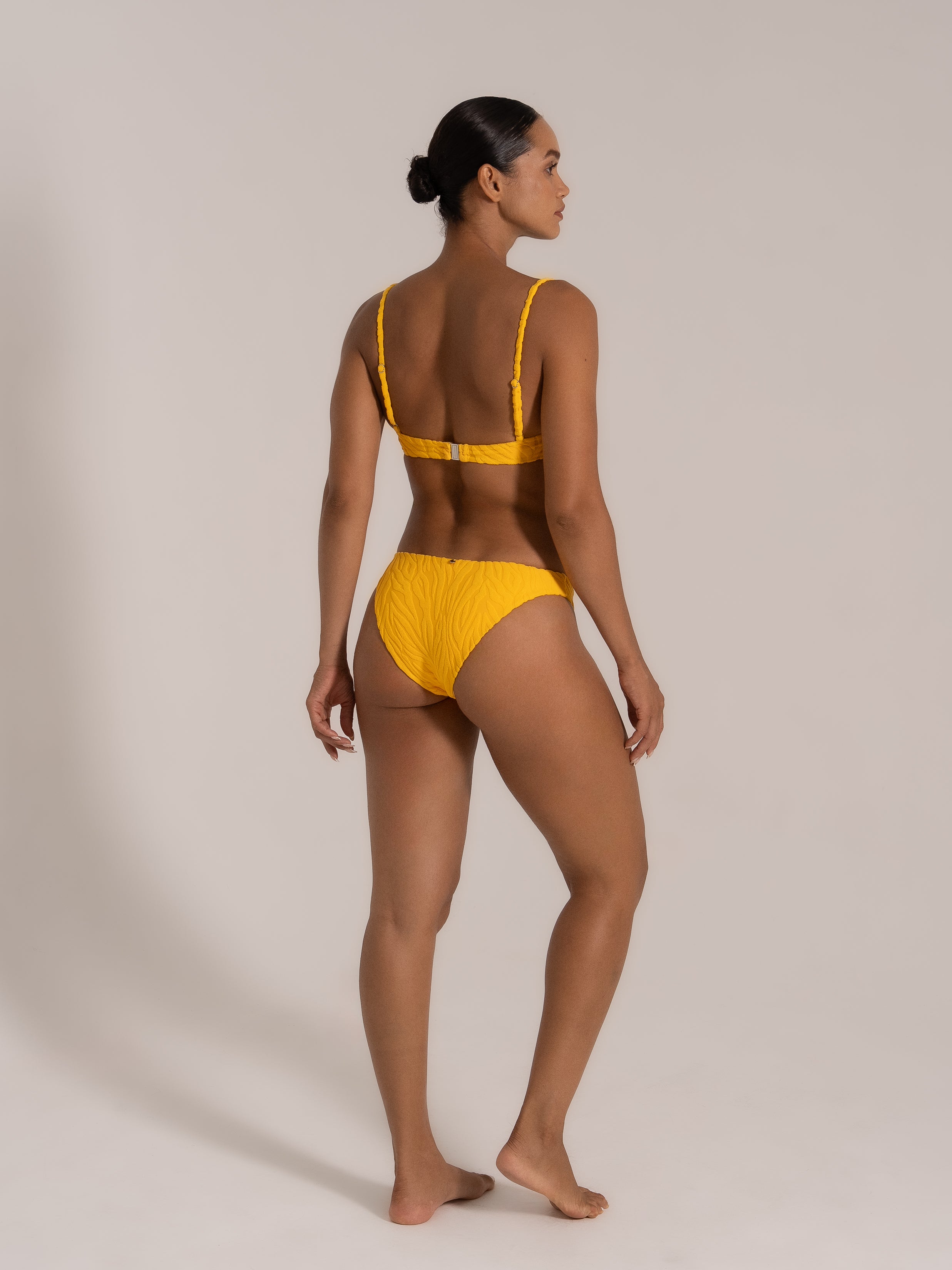 Model showing an angled back view in a yellow textured bikini set with supportive straps and cheeky bottom.